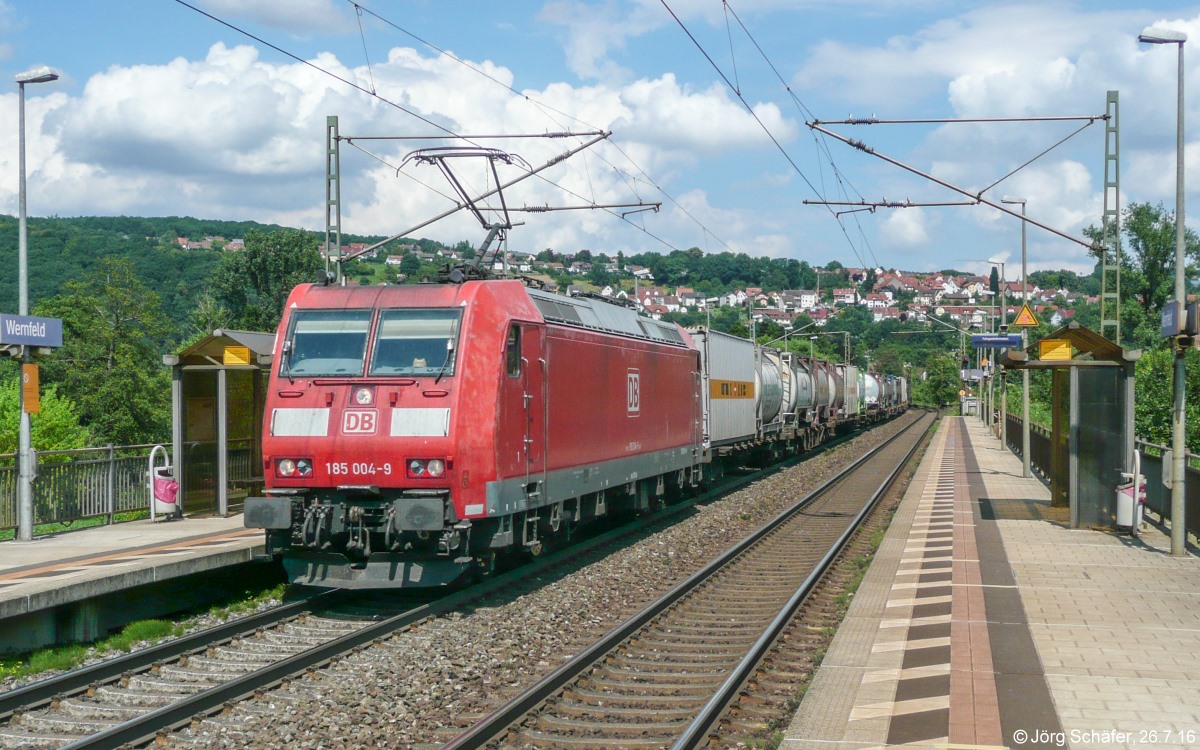 185 004 fuhr am 26.7.16 mit einen Güterzug Richtung Süden durch den neuen Haltepunkt Wernfeld. Im Hintergrund sieht man Adelsberg am Osthang des Maintals.