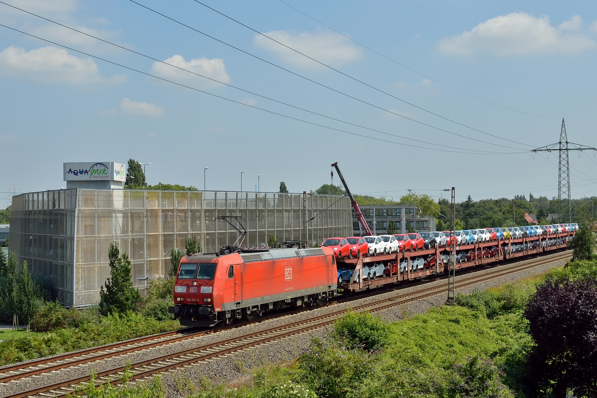 185 051-0 zieht am 31.07.2014 einen Autotransportzug. Die Ladung besteht gänzlich aus Ford KA. Im Hintergrund sieht man die Perle kostenoptimierter moderner Profanbauarchitektur: Das Parkhaus des Aquapark Oberhausen (Spaßbad).