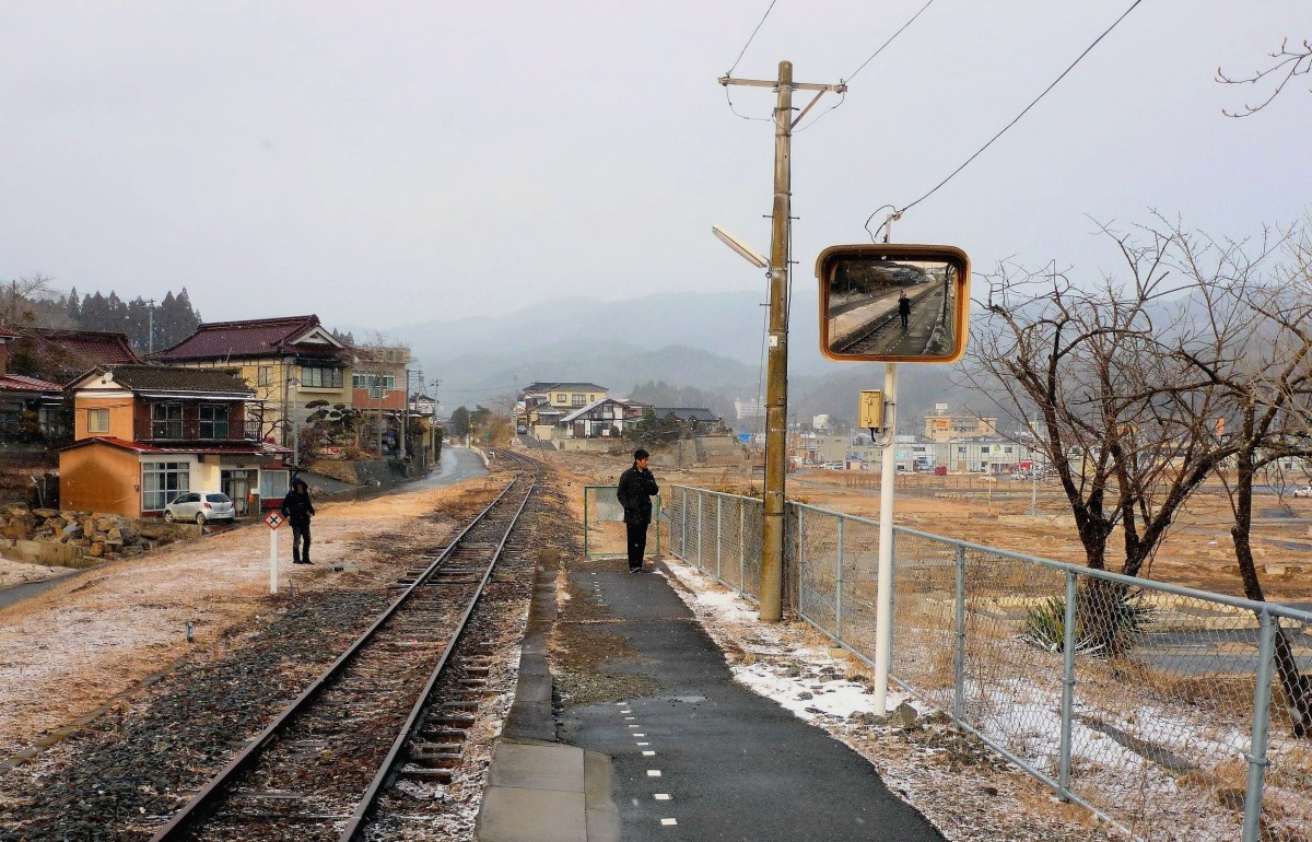 Ôfunato-Linie, Bahnhof Shishiori Karakuwa am 20.Februar 2013: Der Tsunami vom 11.März 2011 hat die ganze Ortschaft hinweggespült; nach dem Einsturz von Oeltanks am Hafen hatte sich der Tsunami als brennende Walze über Shishiori Karakuwa ergossen. Im Hintergrund rechts stehen ein paar Baracken für die Nothilfe. Im Spiegel wird sich der Triebwagen vom oberen Bild nie mehr spiegeln. 