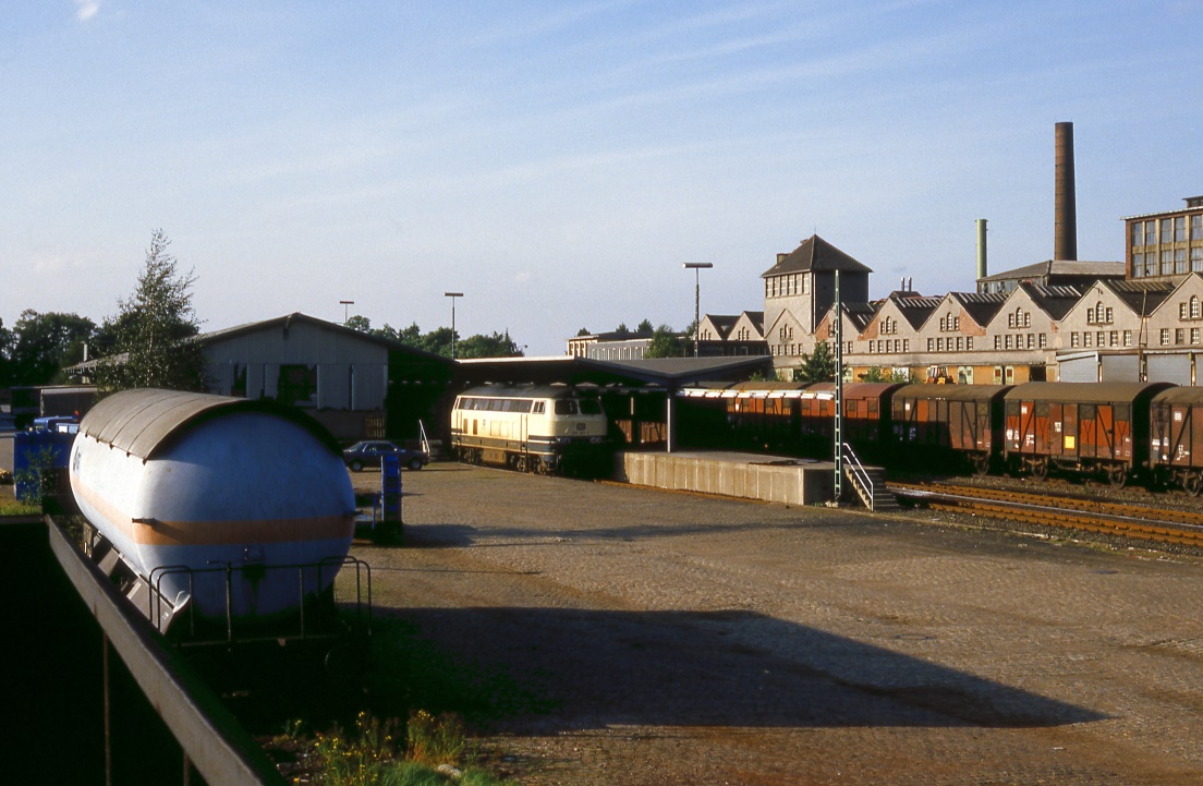 215 021 an der Güterrampe des Bf.Velbert, 02.09.1985. Nachdem die Schnellstraße Velbert - Wuppertal eröffnet worden war, wurde die Stückgutverladung nach Wuppertal Langerfeld verlegt. Die beiden Zugpaare sind entfallen, der Bahnhof wurde zurückgebaut. Anstelle der Gießerei im Hintergrund gibt es nun einen riesigen Baumarkt, der auch große Teile des Bahngeländes nutzt.