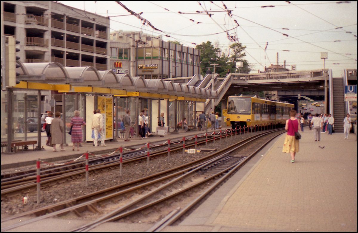 Als die Stuttgarter Stadtbahn noch jung war -

Ein ganz anderes Bild als heute bot die Haltestelle Wilhelmsplatz in Bad Cannstatt. Durch den Mischbetrieb mit den Straßenbahnen der Linie 2 waren noch keine Hochbahnsteige möglich. Erst mit der ursprünglich gar nicht vorgesehenen Umstellung des 2er auf Stadtbahnbetrieb, war der Bau von Hochbahnsteigen möglich. Dabei wurde der gesamte Haltestellenbereich mit einem Glasdach überdeckt, das auch über den Fußgängersteg hinweg führt. Erst kürzlich wurden dann die Bahnsteige für 80 Meter lange Züge verlängert, erst mal zur Entfluchtung des Cannstatter Wasens, später dann für die Doppeltraktion auf der Linie U1.

Wie die Haltestelle 1979 aussah zeigt diese Aufnahme von Kurt:

https://www.bahnbilder.de/bild/deutschland~strassenbahn~stuttgart-keine-stadtbahn/654099/stuttgart-ssb-sl-2-dot4-925.html

Dem Zustand ab 2002 zeigt diese Aufnahme, die Bahnsteige waren noch nicht verlängert:

https://www.bahnbilder.de/bild/deutschland~stadtbahnen-und-u-bahnen~stadtbahn-stuttgart-haltestellen-und-strecken/956400/-zweistufiger-wandel---seit-der.html

Scan vom Farbdia, Ende der 1980iger Jahre 