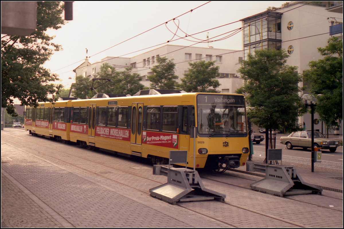 Als die Stuttgarter Stadtbahn noch jung war -

Die Endhaltestelle 'Fellbach Lutherkirche' befindet sich an der selben Stelle wie früher auch die Straßenbahnhaltestelle. Die langgezogene Streifenstrecke kam aus dem Straßenzug, der sich im Bild hinter der Fahrerkabine befindet, rechts außerhalb des Bildes fuhren dann die Straßenbahn ein einer anderen Straße wieder zurück in Richtung Stuttgart.
Aus städtebaulichen Gründen wurde hier auf Hochbahnsteige verzichtet, wobei gerade an Endhaltestellen größerer Fahrgastwechsel herrscht zumal hier auf anschließende Busse umgestiegen werden kann. Erst 2007 wurden hier dann doch Hochbahnsteige gebaut. Schwierig gestaltet sich hier aber die Planung auf eine Verlängerung auf 80 Meter, da sie dann eine wichtige Fußgängerquerung unterbrochen würde.

Ende der 1990 Jahre wurden Planungen recht konkret die U1 in meine Wohngemeinde Kernen im Remstal weiterzuführen. Dazu hätte man ab hier in einem Tunnel unter Fellbach geradeaus nach Osten fahren müssen, um dann auf einer Überlandstrecke Rommelshausen zu erreichen, hier wäre man oberirdische durch Rommelshausen gefahren, wohl auf eingleisiger Strecke um dann in einer über Berg und Tal führenden Abschnitt Stetten im Remstal angefahren hätte. Beide Orte zusammen bilden Kernen im Remstal. Die Trasse wird aber inzwischen leider nicht mehr freigehalten, wo sich die erste Haltestelle in Rommelshausen hätte gebaut werden sollen steht jetzt ein Einfamilienhaus. Da aber ein kleines man kann sagen Dorf für Menschen mit geistiger Behinderung (mein früherer Arbeitsplatz) zwischen beiden Ortsteilen aber aufgegeben wurde und dort ein großes, dicht bebautes Wohngebiet entstehen soll, könnte ich mir durchaus vorstellen, dass die Pläne in geänderter Form eines Tages auch wieder ausgegraben werden. Mich würde es freuen!

Scan vom Farbnegativ, Ende der 1980iger Jahre
