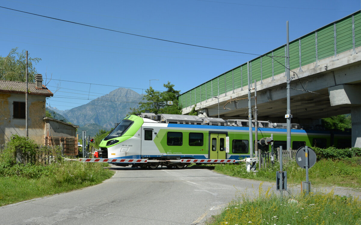 Am 11. Juni 2022 passiert der ETR 104 030 vorschriftsmäßig gesichert den BÜ beim großen Straßenverkehrskreuz Valchiavenna (Sp 36) / Valtellina (Sp 38) im Gewerbegebiet von Colico und fährt weiter in Richtung Sondrio und Tirano im Veltliner Tal.