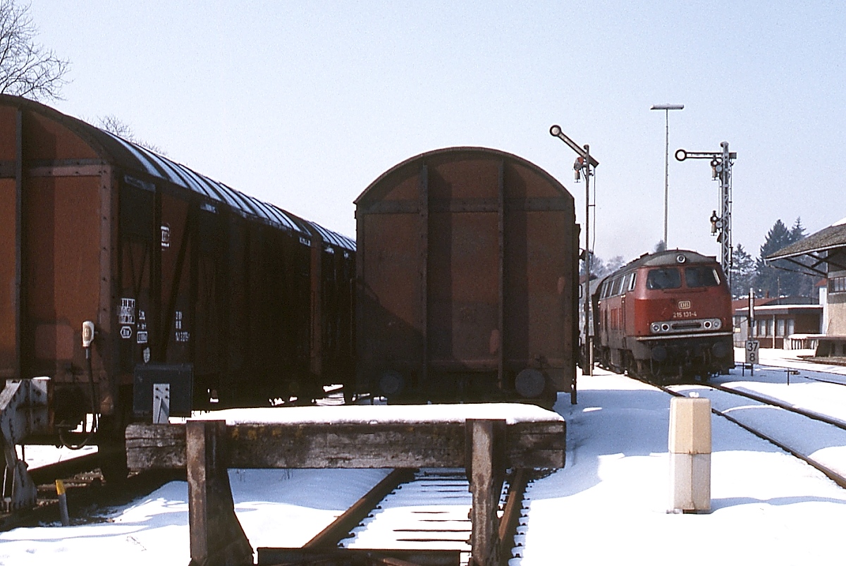 Am 14.02.1983 verlässt 215 131-4 mit einem Nahverkehrszug den Bahnhof Bad Waldsee an der Württembergischen Allgäubahn. Zwar drängen sich die Güterwagen etwas in den Vordergrund, allerdings sind sie ein Beleg dafür, dass es damals noch ordentlichen Güterverkehr gab, heute befindet sich hier ein Parkplatz.