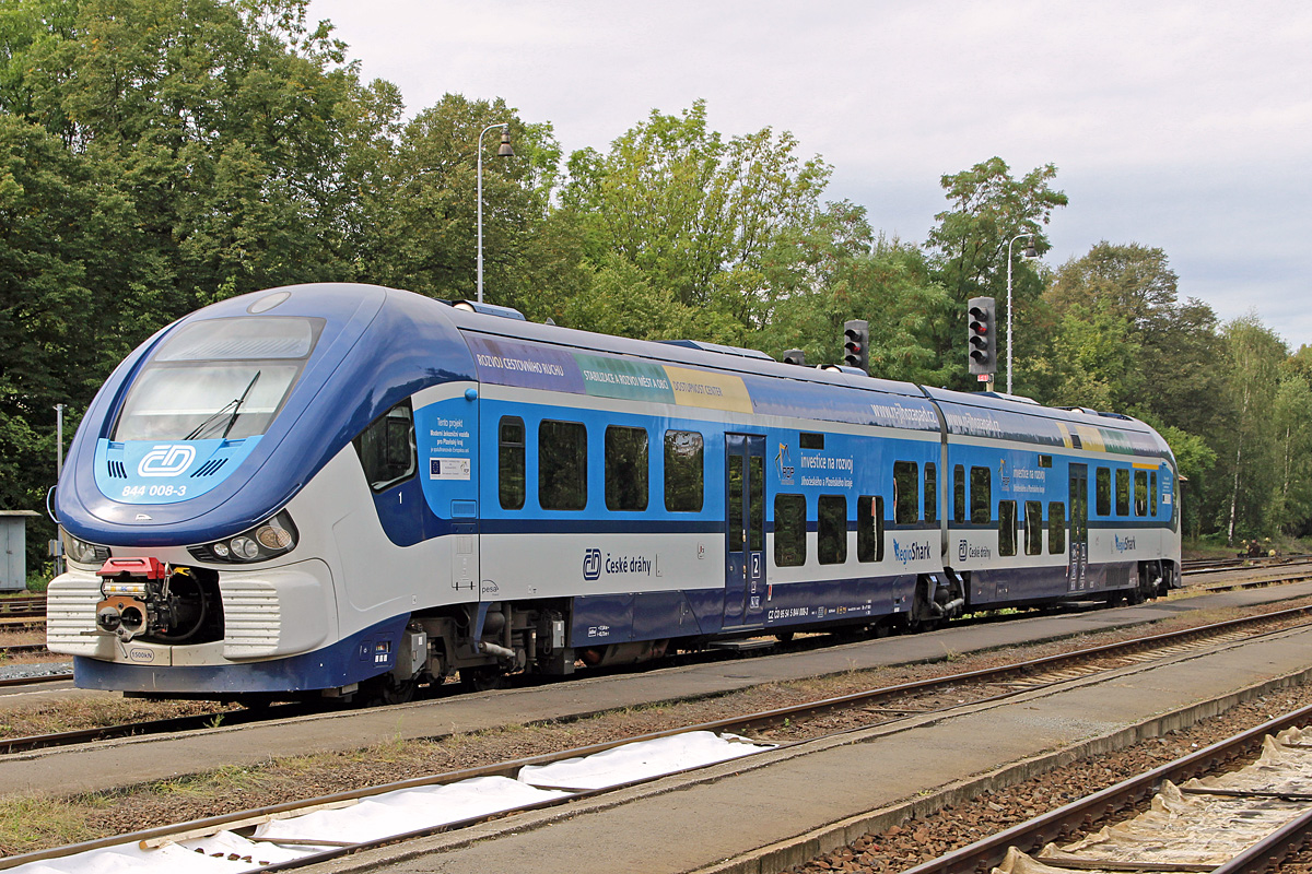 Am 15.09.2015 wartet 844 008 in Domažlice auf seinen nächsten Einsatz als Os nach Plzeň. Die in Polen gebauten RegioSharks sind eine interessante Bereicherung des tschechischen Fahrzeugparks. Auch der Fahrkomfort und die Ausstattung können überzeugen, die Triebzüge werden ja nur im Nahverkehr verwendet. Ein bemerkenswertes Detail sind die seitlichen Fensterscheiben, die nicht senkrecht eingebaut sind. Deren oberes Ende ist etwas weiter außen als das untere Ende, den Effekt sieht man, wenn es regnet, man hat (fast) immer klare Sicht nach draußen. 