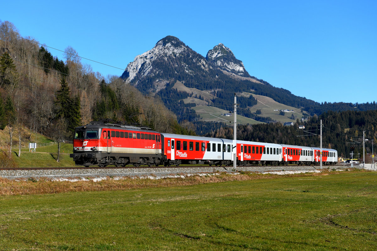Am 17. November 2018 war eine zweite 1142 mit  Pflatsch  auf der Salzkammergutbahn unterwegs. Bei Tauplitz konnte ich die 1142.685 vor dem REX 4413 nach Attnang-Puchheim an der Haltestelle Kulm aufnehmen. Diese wird nur genutzt, wenn Skisprung-Wettbewerbe stattfinden. Im Hintergrund sieht man den Gwendlingstein und den Hechlstein. Die abgebildete Lok ist ein Sonderling, weist sie doch zwei verschiedene Frontgestaltungen auf. Auf der Seite des Führerstands 1 hat sie umlaufende Griffstangen und die großen Scheinwerfer in der Ursprungsversion. Am Führerstand 2 wurden die kleinen Standard-Scheinwerfer und die kurzen Griffstangen verbaut, welche viele der Loks im Rahmen von Ausbesserungen in den 1990-er Jahren erhielten. In Deutschland waren solche  Janus-Köpfe  bei den Einheits-E-Loks gang und gäbe, bei den ÖBB Baureihen 1042/1142 eine einmalige Ausnahme. 