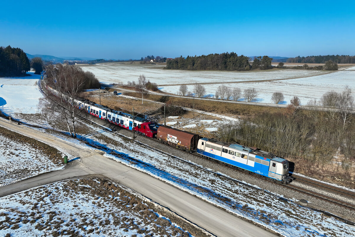 Am 21. Januar 2026 k�ndigten sich bereits die Bauma�nahmen zur Generalsanierung der Strecke zwischen N�rnberg und Regensburg an. Bei Sinngr�n in der Oberpfalz waren etliche Baufahrzeuge zu sehen, die unsch�n im Motiv standen. Daher wurde die Drohne gestartet, um trotzdem an dieser Stelle fotografieren zu k�nnen. Als die hier gezeigte �berf�hrungsfahrt vorbei fuhr, war der ganze Bautrupp jedoch kurz vorher zur Mittagspause aufgebrochen. Zwei Triebwagen des Typs Alstom Coradia Stream wurden nach Rum�nien transportiert. Die dortige Eisenbahngesellschaft AFR hat 20 elektrische Triebz�ge dieses Typs f�r den Fernverkehr beschafft. F�r die �berstellung zum Grenz�bergang nach Passau war die 151 075 von SRI zust�ndig. Der Bundesbahnklassiker befand sich nach Verkauf durch DB Cargo einst im Leasing-Pool von Railpool. Daf�r hatte die Lok im Jahre 2020 auch eine interessante Lackierung erhalten, bei welcher die Farbgebung vom urspr�nglichen ozeanblau-beige zum silbernen Railpool Farbton hin wechselte. �ber die Zeit hat das �u�ere der Lok sichtbar gelitten. Der neue Eigner SRI hat dann der Maschine auch wenige Tage nach Entstehen dieser Aufnahme eine Auffrischung spendiert. Der silberne Lack am F�hrerstand 1 verschwand. Nun zeigt sich 151 075 auch dort wieder in ozeanblau-beiger Farbgebung mit Zierstreifen.  