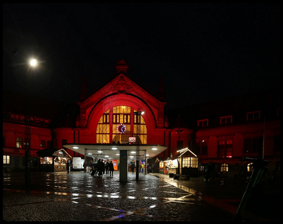 Am 23.11.2021 hielt ich bei Einbruch der Dunkelheit die illuminierte Fassade des Hauptbahnhof Osnabrück im Bild fest. Zukünftig soll der Bahnhof zur Vorweihnachtszeit regelmäßig in dieser Form angestrahlt werden, wobei sich die Farben immer wieder ändern.
Aus dieser Perspektive ahnt man nicht, dass es sich um einen Turmbahnhof handelt und sich hinter dem Gebäude zwei Bahnstrecken kreuzen.