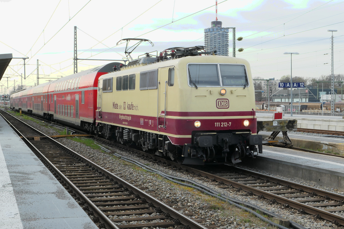 Am 26. März 2024 bespannte die 111 212 der DB-Gebrauchtzug in ihrem auffälligen Tee-farbenen Anstrich den RE16 Augsburg - Nürnberg. Das markante Gebäude hinter der Lokomotive wurde in den 1970er-Jahren als  Augsburger Maiskolben  bezeichnet.
Augsburg Hbf, 26. März 2024 