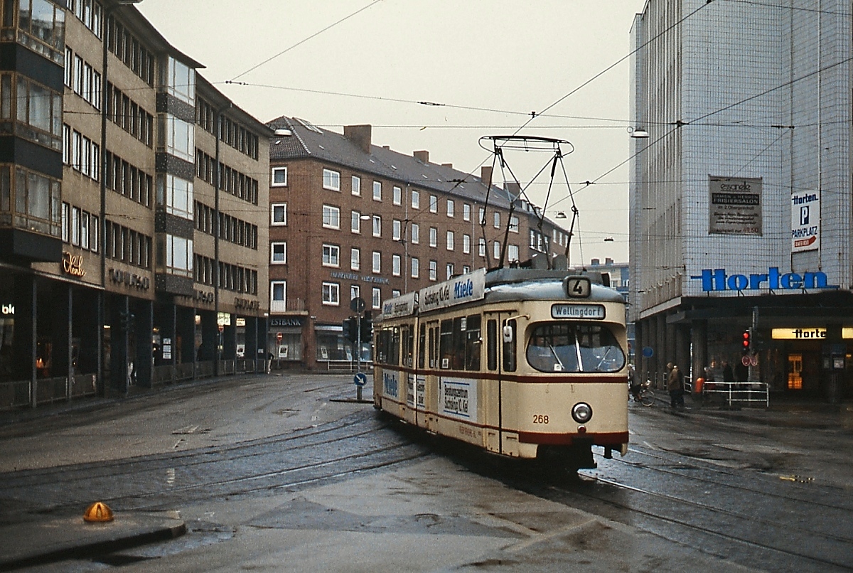 Am 27.04.1985 überquert der Kieler Tw 268 auf dem Weg nach Wellingdorf den Dreiecksplatz. Seit 30.09.1984 bogen die Straßenbahnen wegen der schon laufenden Bauarbeiten für den Busverkehr nicht mehr über die links abbiegenden Gleise in die Bergstraße ab, sondern befuhren die sogenannte  Katastrophenschleife  durch die Brunswiker Straße.