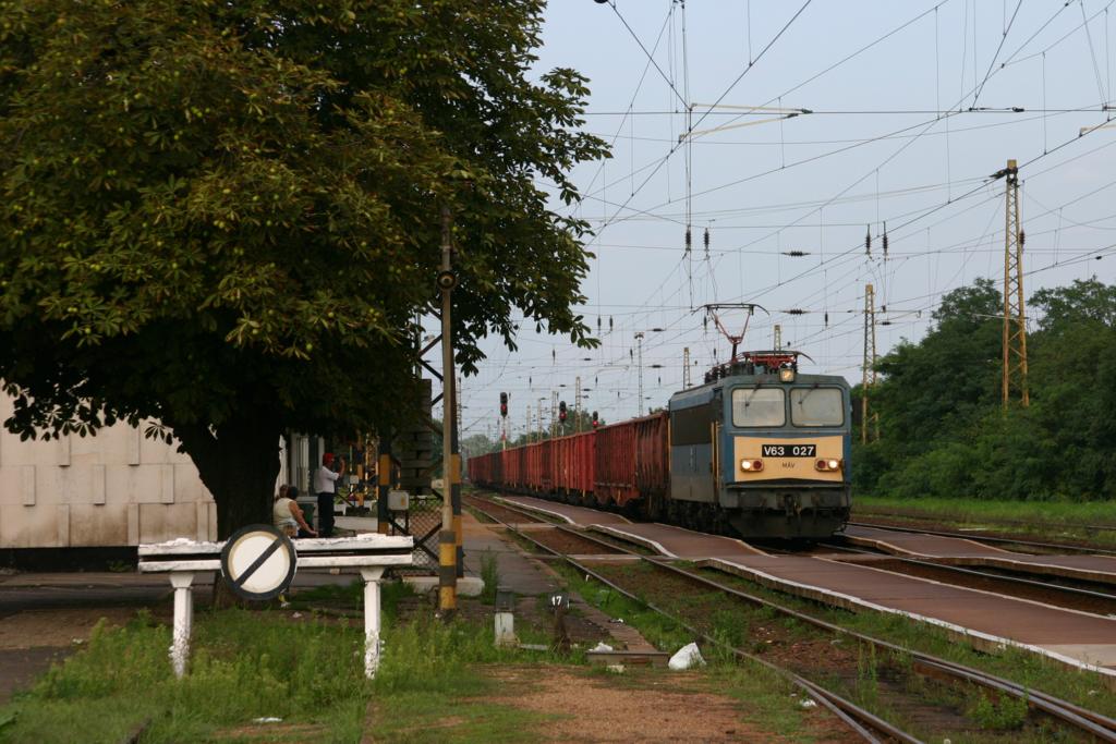 Am 29.08.2005 fährt V 63027 mit einem beladenen Kohlezug aus Richtung Miskolc kommend durch den Bahnhof Mezökövesd. Wie auch in Ungarn üblich, steht der Fahr-
dienstleiter mit Mütze vor dem Bahnhof Gebäude und grüßt bei der Durchfahrt.