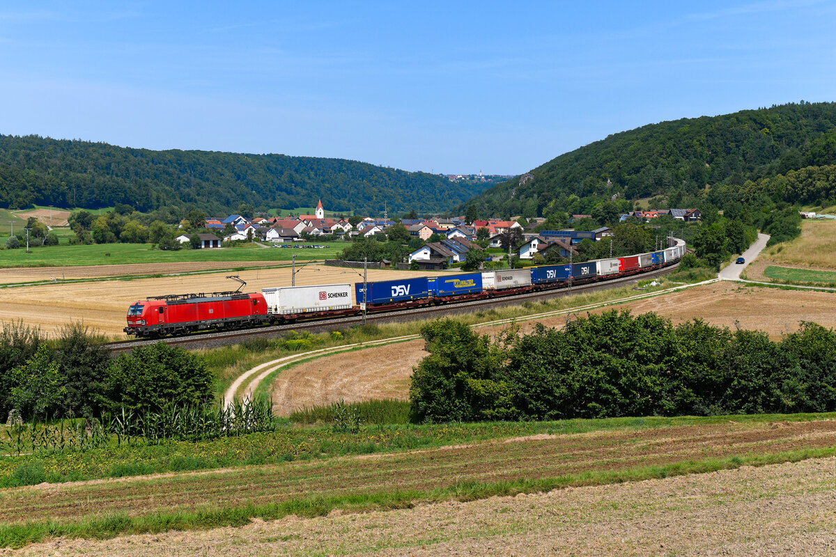 Am 31. Juli 2024 bespannte die 193 328 den KLV-Zug KT 42152 von Verona Q.E. nach Rostock Seehafen. Die Beladung des Zuges wies schon sinnbildlich auf ein zum Aufnahmezeitpunkt kurz bevorstehendes Ereignis hin. Das dänische Transportunternehmen DSV übernahm die Logistik-Tochter der Deutschen Bahn, DB Schenker, für 14,3 Milliarden Euro. Mit dem erzielten Erlös soll die Bahn einen großen Teil ihres Schuldenbergs abbauen. Ob es allerdings tatsächlich auf lange Sicht klug war, den größten Gewinnbringer im Konzern abzustoßen, wird die Zukunft zeigen. Die hier gezeigte Aufnahme entstand in Breitenfurt im Altmühltal.