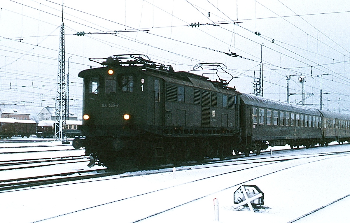 Am 31.12.1977 verlässt die 144 509-7 mit einem Nahverkehrszug den Bahnhof Freilassing in Richtung Berchtesgaden. Die E 44.5 wurden zwischen 1933 und 1935 speziell für diese steigungsreiche Bahnstrecke beschafft und beförderten fast ein halbes Jahrhundert lang die meisten Züge auf dieser Verbindung.