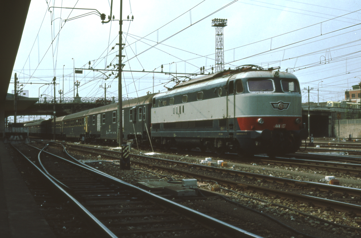 Am Mittag des 12. August 1980 fährt die E444 107 mit dem Rapido Milano C - Napoli C aus dem Bahnhof Bologna Centrale aus. Bis auf den Speisewagen besteht der Zug ausschließlich aus Waggons des Typs UIC-X in der neuesten Ausführung, in der Farbe  Grigio Adesia , einem dunklen Grau. Canon AE1, Canoscan, GIMP - erst mit der damals hochmodernen Canon AE1, die ich im Herbst 1979 gekauft habe, kam bei mir die Fotografie so richtig in Gang.