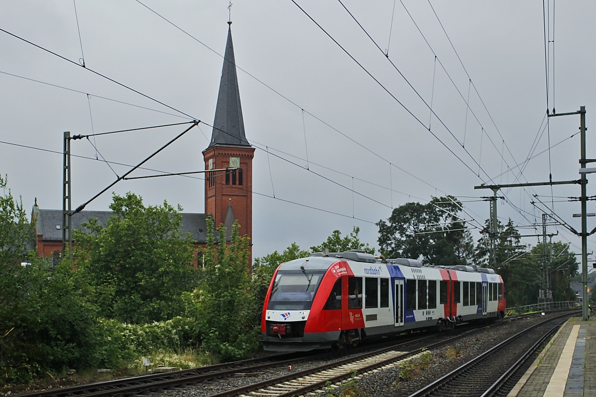 Am trüben Vormittag des 10.07.2023 trifft ein Nordbahn-LINT aus Bad Oldesloe in Neumünster ein, im Hintergrund die Kirche Sankt Maria-Sankt Vicelin