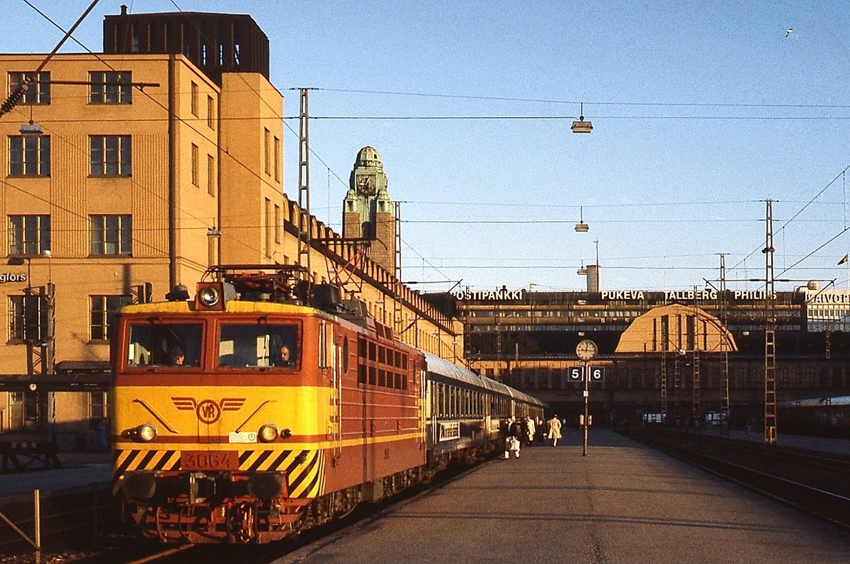 An einem Maiabend 1988 steht die Sr1 3064 um 21:15 Uhr vor einem Nachtschnellzug im Bahnhof von Helsinki, der damals noch keine Bahnsteigüberdachung hatte. Mit der Elektrifizierung begannen die VR erst in den späten 1960er Jahre und so sind die 112 bei Nowotscherkassk/Sowjetunion ab 1973 beschafften Sr1 die ersten elektrischen Staatsbahnlokomotiven in Finnland.