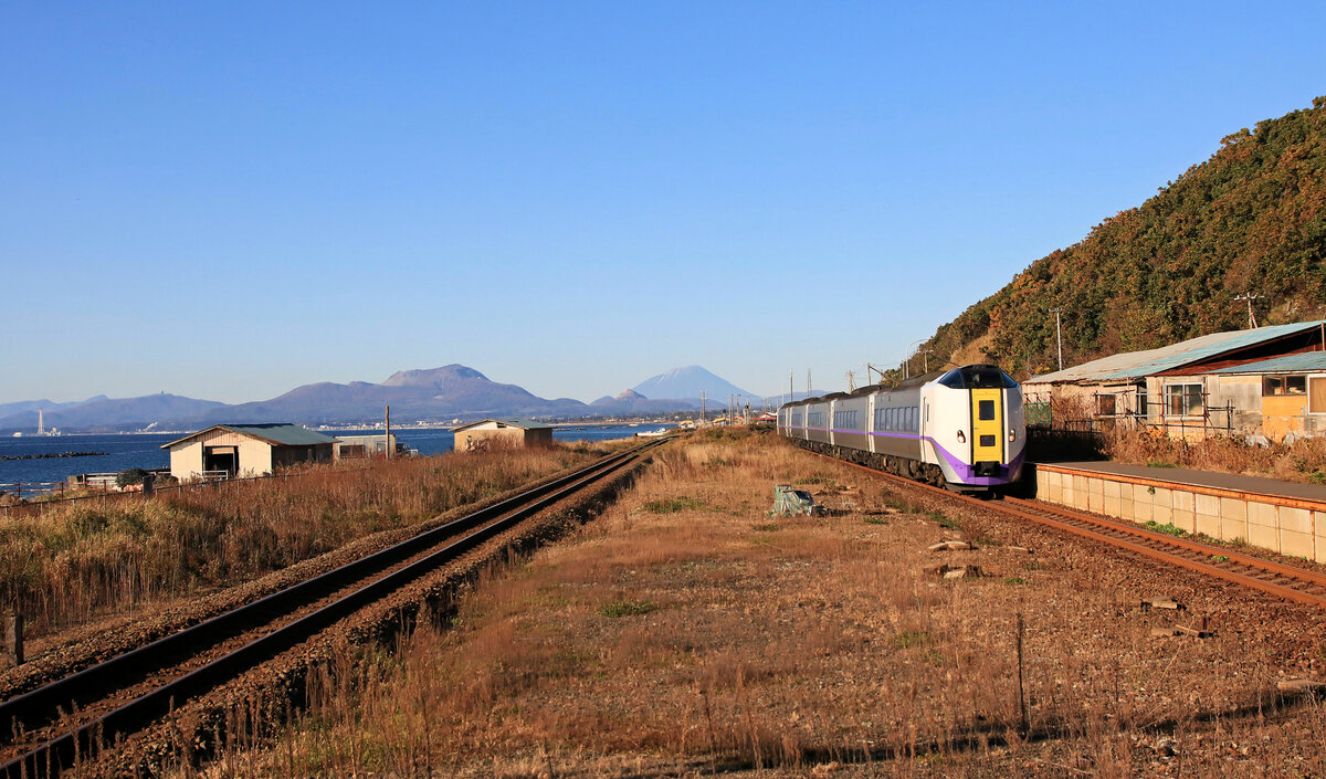 An der Hauptlinie, die die vorübergehende Endstation des Shinkansen in Hakodate und die Hauptstadt von Hokkaidô, Sapporo, verbindet: Durchfahrt eines Diesel-Expresszuges der Serie KIHA 260/261-1000. Kogane, 26.Oktober 2022. Im Hintergrund die drei Vulkane Usu-zan, der kleine Shôwa Shinzan, und in der Ferne der mächtige Yôteizan.  