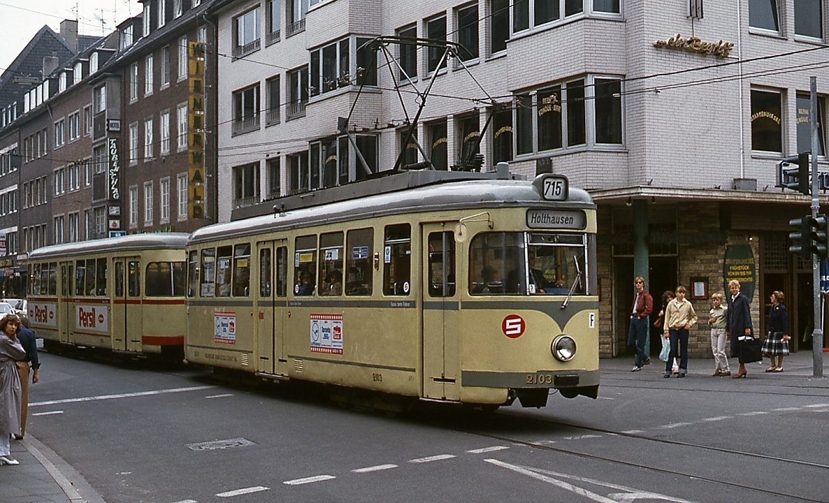 Anfang der 1980er Jahre fährt Tw 2103 auf der Hunsrückenstraße in Richtung Holthausen. Nach der Gründung des Verkehrsverbundes Rhein-Ruhr erhielten die Linien im Verbund zur Vermeidung von Doppelnummern dreistellige Linienbezeichnungen, in Düsseldorf wurde den Straßenbahnlinien eine  7  vorgestellt und so wurde aus der  15  die  715 . Der 2103 besitzt noch die bis Mitte der 1970er Jahre verwendete graue Zierlinie der Stadtlinien, der rote Streifen war bis dahin den Fernbahnlinien vorbehalten. Bei dem Beiwagen handelt es sich um einen früheren Triebwagen.