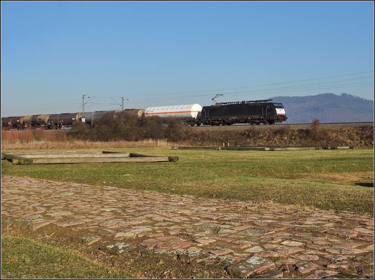 Antike und heute. Die badische Hauptbahn folgt in weiten Bereichen den alten Römerstraßen. Hier in Friesenheim wird ein Stück Römerstraße mit Raststätte an einer antiken Kreuzung gepflegt, netterweise direkt neben der Bahnstrecke. 189 090 mit einem Güterzug auf Römerpfaden. Im Hintergrund ist noch der <I>Abnuba mons</I> zu sehen, zu deutsch Schwarzwald. Februar 2019.