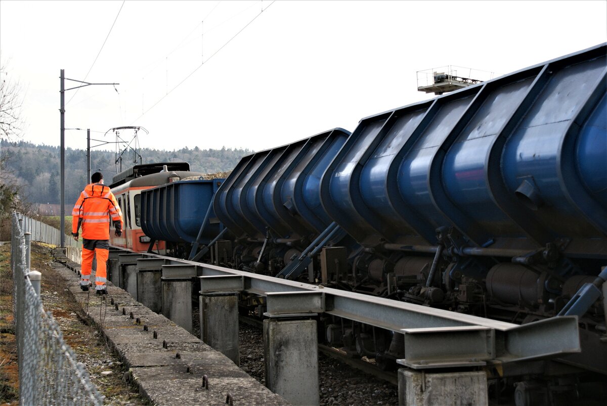 ASM BTI Aare Seeland mobil AG Biel-Täuffelen-Ins-Bahn: Von vorne nach hinten Fa 576 + Fa 571 + Fa 572 + Be 4/4 302, Entladen von Zug 8281 (eine Sache von einigen Sekunden!), fernbedient aus dem hinteren Führerstand des Be 4/4 304, Sutz, 9. Februar 2021. Seitlich unter den Stahlträger ausgefahrene Querstangen verhindern ein Abstürzen der Wagen beim Abkippen der Ladung (erkennbar beim vordersten Betonsockel, sowie beim dritten solchen von rechts). Die pneumatischen Einseitenkastenkipper wurden von der Lausitzer Bergbau-Verwaltungsgesellschaft mbH erworben, im Dampflokwerk Meiningen der Deutschen Bahn AG aufgearbeitet, dort auch von 900 mm auf 1000 mm umgespurt und von der Werkstätte Täuffelen ab 1998 fertig ausgerüstet. Nach nicht nachgeprüften Angaben wurden diese Abraumwagen 1964 von der Waggonfabrik Arad in Rumänien nach Plänen des VEB Waggonbau Bautzen (Type TGL 5834 25/900) gebaut und waren im Braunkohletagebau Berzdorf bei Görlitz eingesetzt.