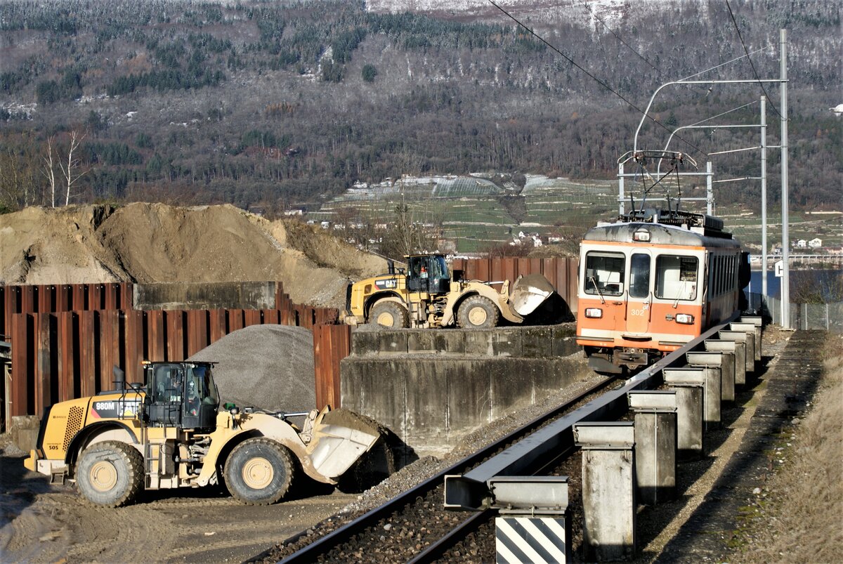 ASM BTI Aare Seeland mobil AG Biel-Täuffelen-Ins-Bahn: Nach Erschöpfung der Grube Finsterhennen und Kündigung des Transportauftrages verkehrten am 10. Dezember 2021 nach 18 Jahren ein letztes Mal die Zugpaare 8233/8238 und 8245/8250 für die Hurni Kies- und Betonwerk AG in Sutz. Inzwischen wurde das Anschlussgleis in Sutz gesperrt und die Fahrleitung dort abgebaut. Das Bild vom 7. Dezember 2021 gewährt nochmals einen Blick  über den Zaun  in Sutz auf Einrichtungen und Arbeitsweise. Zug 8245 hat seine Ladung Kies in die Senke gekippt und danach vorgezogen. Während ein Pneulader darauf wartet, unter ausgeschalteter und geerdeter Fahrleitung die Kippwagen mit Aushub und Bauschutt für die Rückfahrt zu beladen, räumt ein zweiter Pneulader das angelieferte Kies weg. Die Stahlkonstruktion auf Betonsockeln diente der Absturzsicherung der Kippwagen. Diese besitzen unter die Stahlträger pneumatisch ausfahrbare Querstangen. Zuletzt verkehrte während mehr als einem Jahr stets die gleiche Komposition: Be 4/4 302 (orange) + Fa 572 + Fa 571 + Fa 576 + Be 304 (grau). 