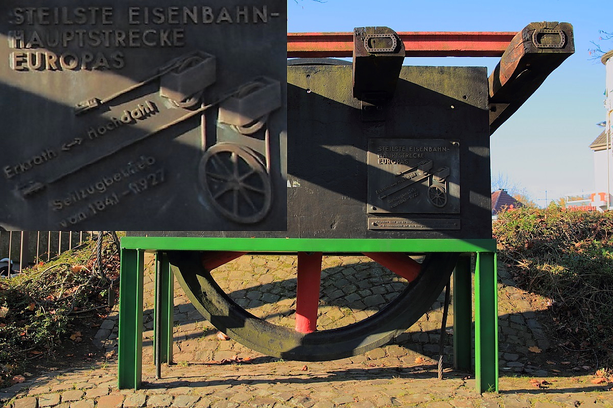 Auf der Bahnlinie von Erkrath nach Hochdahl muss auf 2,5 km Länge ein Höhenunterschied von 82 m überwunden werden. Als die Düsseldorf-Elberfelder Eisenbahngesellschaft die Steilrampe 1841 in Betrieb nahm, wurden die Züge zunächst von einer stationären Dampfmaschine in Hochdahl an einem Seil bergwärts gezogen. Schon wenig später löste ein System, bei dem die talwärts fahrenden Züge die bergwärts fahrenden mithilfe eines Seils, das über am Ende der Steilrampe installierte Umlenkrollen lief, hinauf nach Hochdahl zogen, die Dampfmaschine ab. An diesen Betrieb erinnert ein Denkmal mit der Umlenkrolle in Hochdahl, das Verfahren ist auf dem vergrößerten Bildausschnitt der Gedenktafel (links oben) zu erkennen (Aufnahme vom 01.11.2015). Erst 1927 wurde dieser umständliche Betrieb durch Vorspann- und Schubloks abgelöst. 