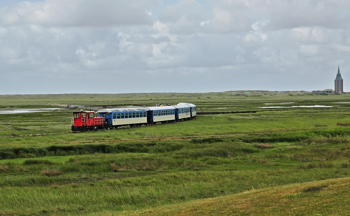 Auf dem Weg vom Anleger nach Wangerooge Ort hat 399 107-2 am 22.06.2018 die Westlagune durchquert, im Vordergrund die selten befahrene Weststrecke der Inselbahn. Im Hintergrund der Neue Westturm von 1932, ursprünglich Leuchtturm, heute dient er als Jugendherberge.