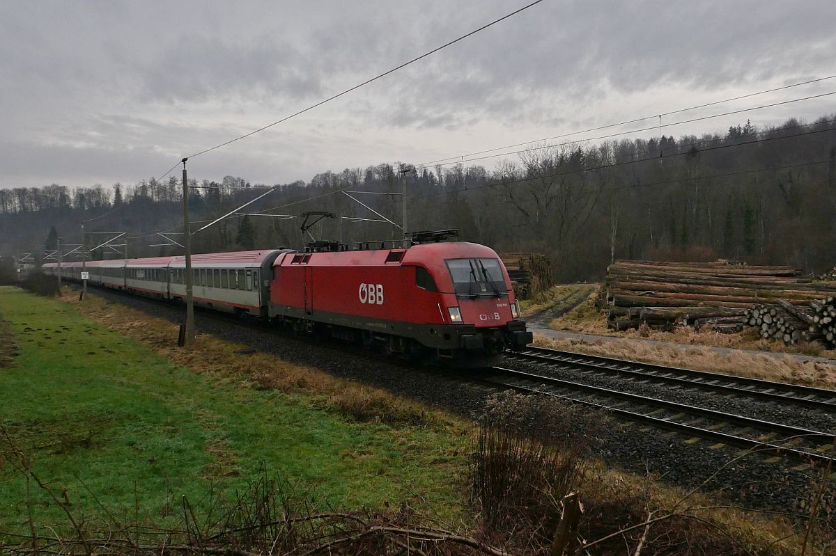 Auf der Fahrt von Innsbruck nach Bochum zieht 1016 013-5 die Wagen des IC 118  BODENSEE  im Schussentobel bei Kilometer 165.4 an einem Holzlager vorbei (08.01.2023)