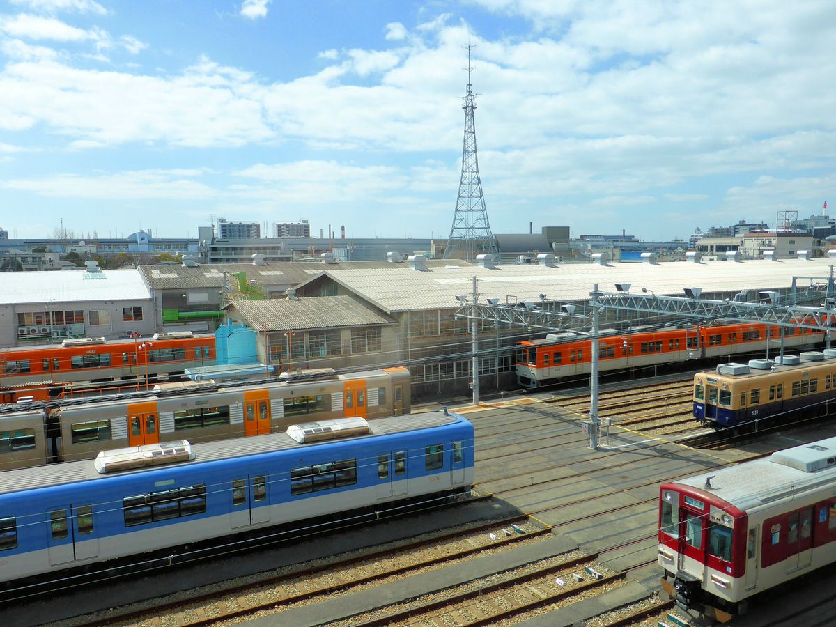 Auf der Fahrt im Zug des Hanshin-Konzern kommt man auch am Betriebswerk Amagasaki vorbei, in dem Hanshin Expresszüge (beige/orange und silbrig mit orangen Türen), Hanshin Lokalzüge (hell- bzw. dunkelblau) und ein Zug des Kintetsu-Konzerns stehen, der aus der Stadt Nara im Osten von Ôsaka gekommen ist. 22.März 2014. 