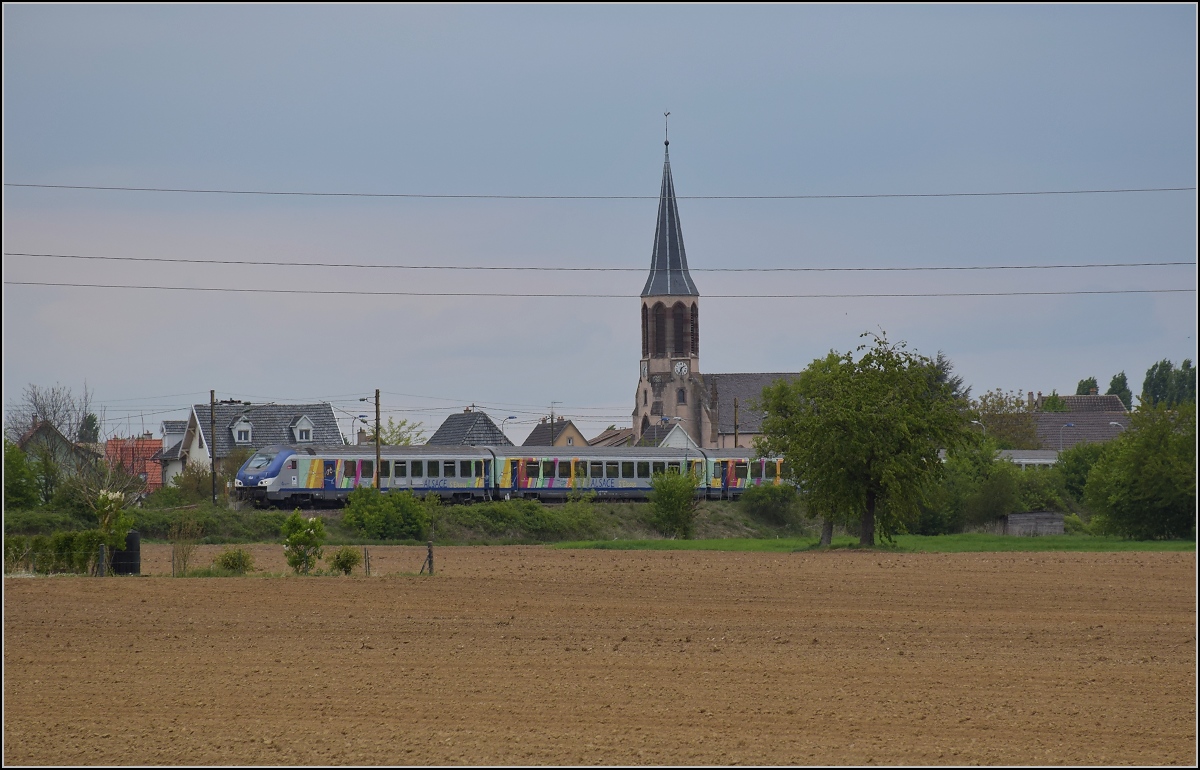 Bahn im Elsass. Bei aufziehendem Schlechtwetter fährt ein TER an Raedersheim vorbei. Vor der markanten Kirche passt auch ein markanter Steuerwagen ins Bild. April 2017.