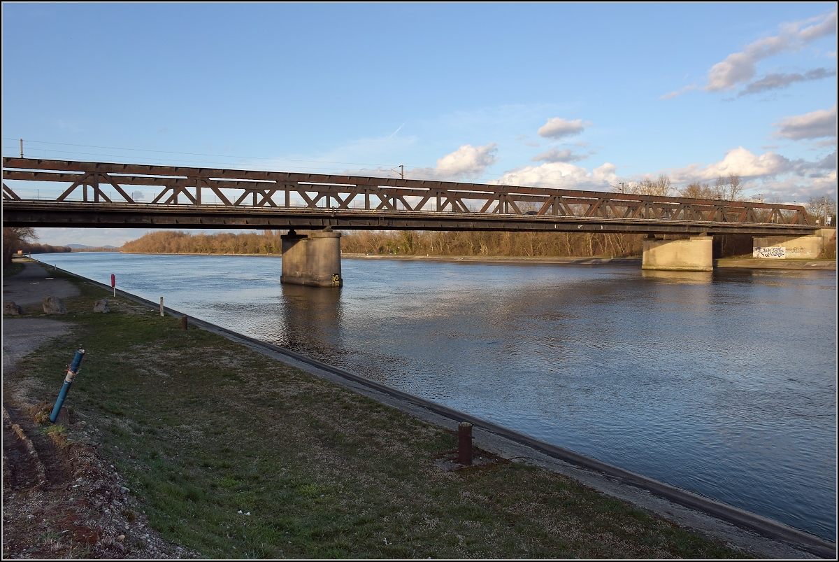 Bahn im Markgräfler Land. Das längere Stück der Verbindungbahn Müllheim - Mühlhausen liegt im Elsass. Hier noch die zweite  Rheinbrücke  über den Rheinseitenkanal, der bereits vollständig im Elsass liegt. Im Hintergrund ist der Kaiserstuhl sichtbar, wo man noch von einer Wiederherstellung des Bahnverkehrs Freiburg - Colmar träumt. Hier weiter südlich ist es bereits Realität, auch dank mittlerweile heute über Kehl geführten TGV-Verbindung nach Freiburg. März 2019.