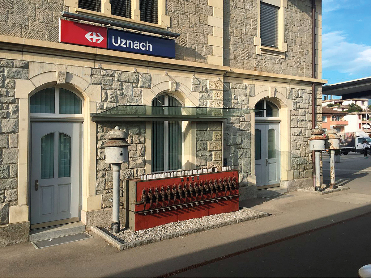 Bahnhof Uznach SG, erhaltenes historisches Stellwerk vor dem Gebäude. 3 ebenfalls historische Bahnhofsglocken ergänzen die Anlage. Aufnahme vom 28. April 2018