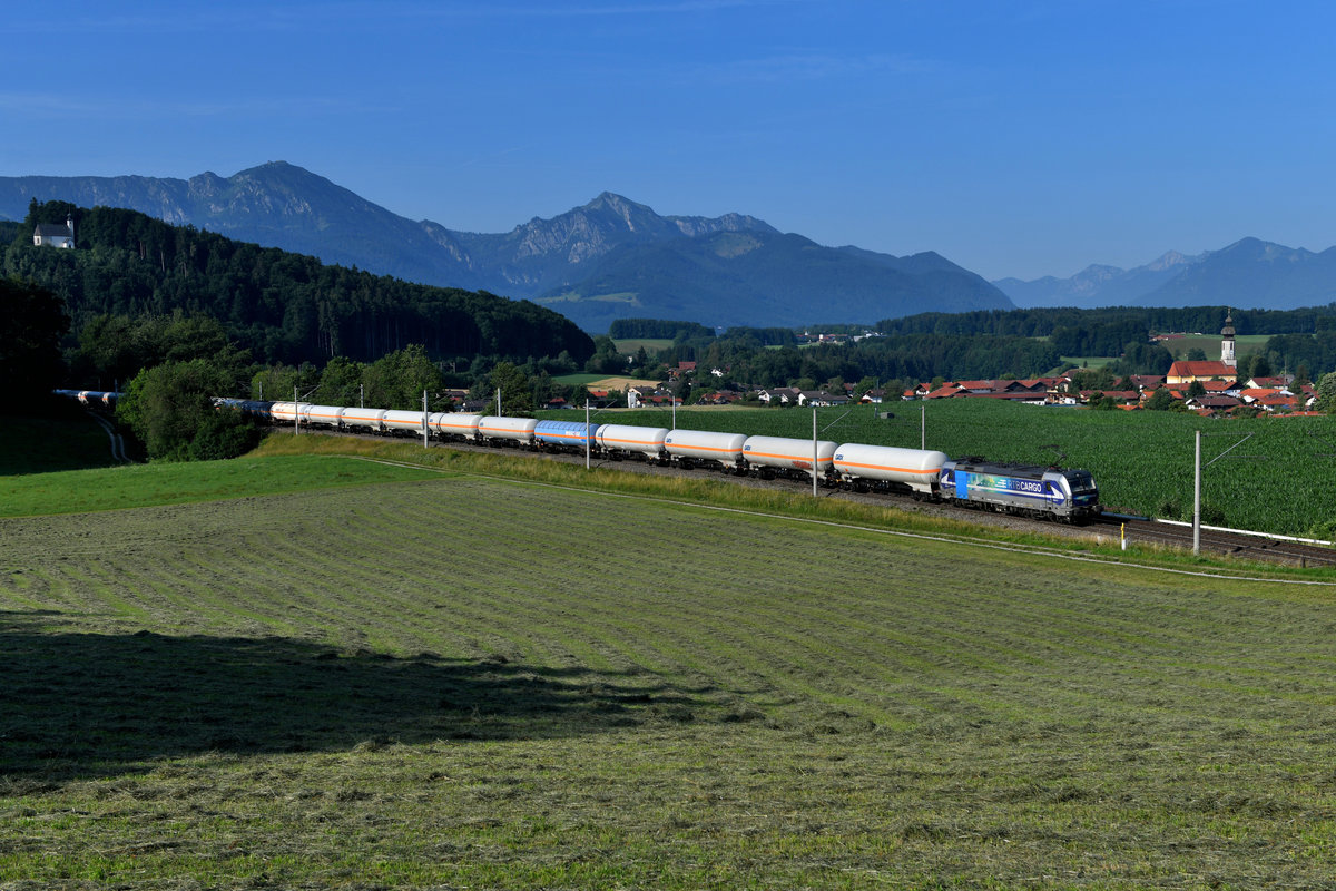 Bei Axdorf findet man dieses schöne Motiv mit Blick auf das St. Georgskircherl, die Chiemgauer Alpen mit Hochgern und Hochfelln, sowie die Ortschaft Vachendorf mit der Kirche Mariä Himmelfahrt. Am 20. Juli 2019 war die Wiese im Vordergrund frisch gemäht worden, dementsprechend war ein Betreten möglich und es roch angenehm nach Heu. Ein passender Zug kam auch zur rechten Zeit. Mit einem aufgrund der Sperre der Passauerbahn umgeleiteten Gaskesselwagenzug nach Hegyeshalom passierte die 193 810 der Rurtalbahn die oberbayerische Bilderbuchlandschaft. Hinweis: Normalerweise kommt bei mir eine Bildmanipulation nicht vor, in diesem Falle habe ich allerdings einige störende Hochspannungsleitungen digital entfernt. 