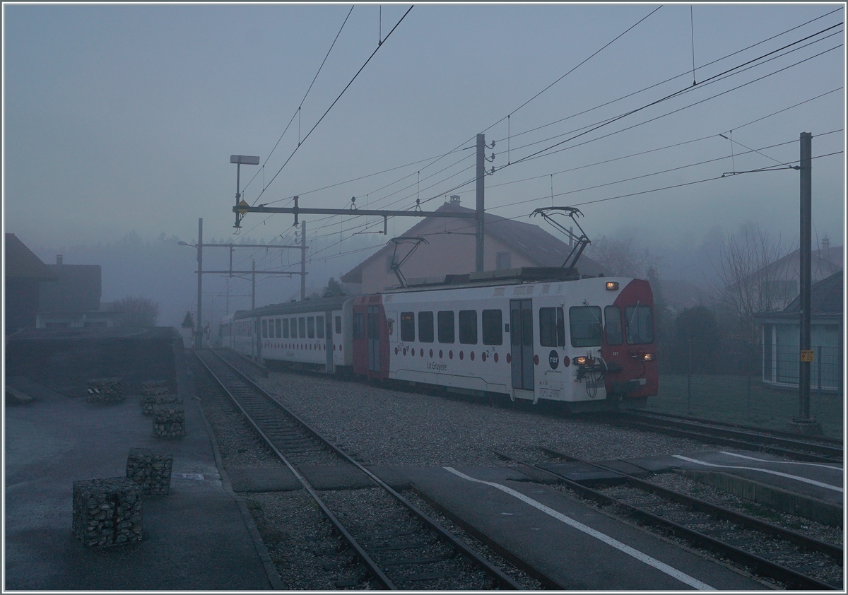 Bei dichtem Nebel und noch in der Morgendämmerung erreicht der TPF Be 4/4 121 mit dem B 207 (ex MOB), B209 (ex MOB) und dem ABt 221 den Bahnhof von Broc Village. Der Zug ist als RER 60 14954 von Bulle nach Broc Farbrique unterwegs. 

26. Nov. 2020