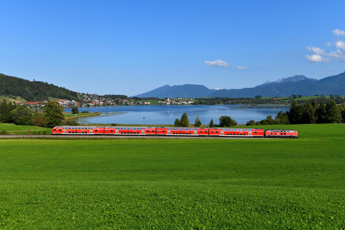 Bei Reinertshof ergibt sich dieser Blick auf den Hopfensee und die Ortschaft Hopfen am See. 218 414 schob am 20. September den RE 57513 nach München HBF. Die Reisenden hatten an diesem schönen Tag sicherlich eine angenehme Fahrt in den Doppelstockwagen. Im Hintergrund erkennt man das Ammergebirge. 