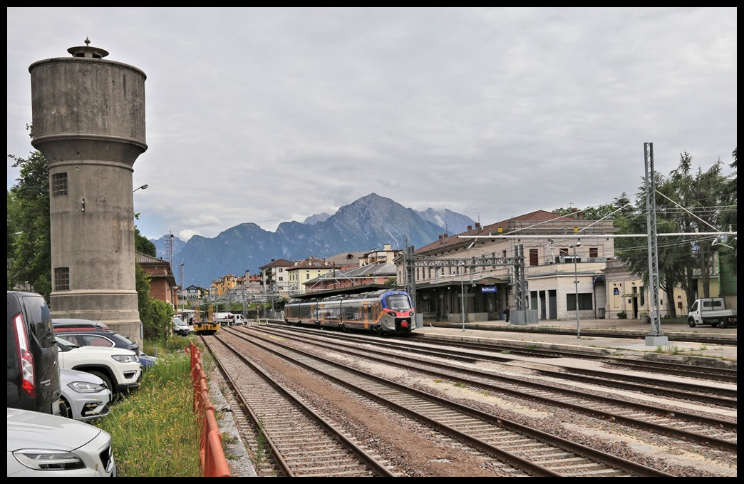 Blick auf den Bahnhof Belluno, dessen alter Wasserturm noch auf der dem Hauptgebäude abgewandten Seite empor ragt. Im Bahnhof steht am 30.5.2022 um 11.00 Uhr ein Elektrotriebzug in Richtung Norden am Bahnsteig.