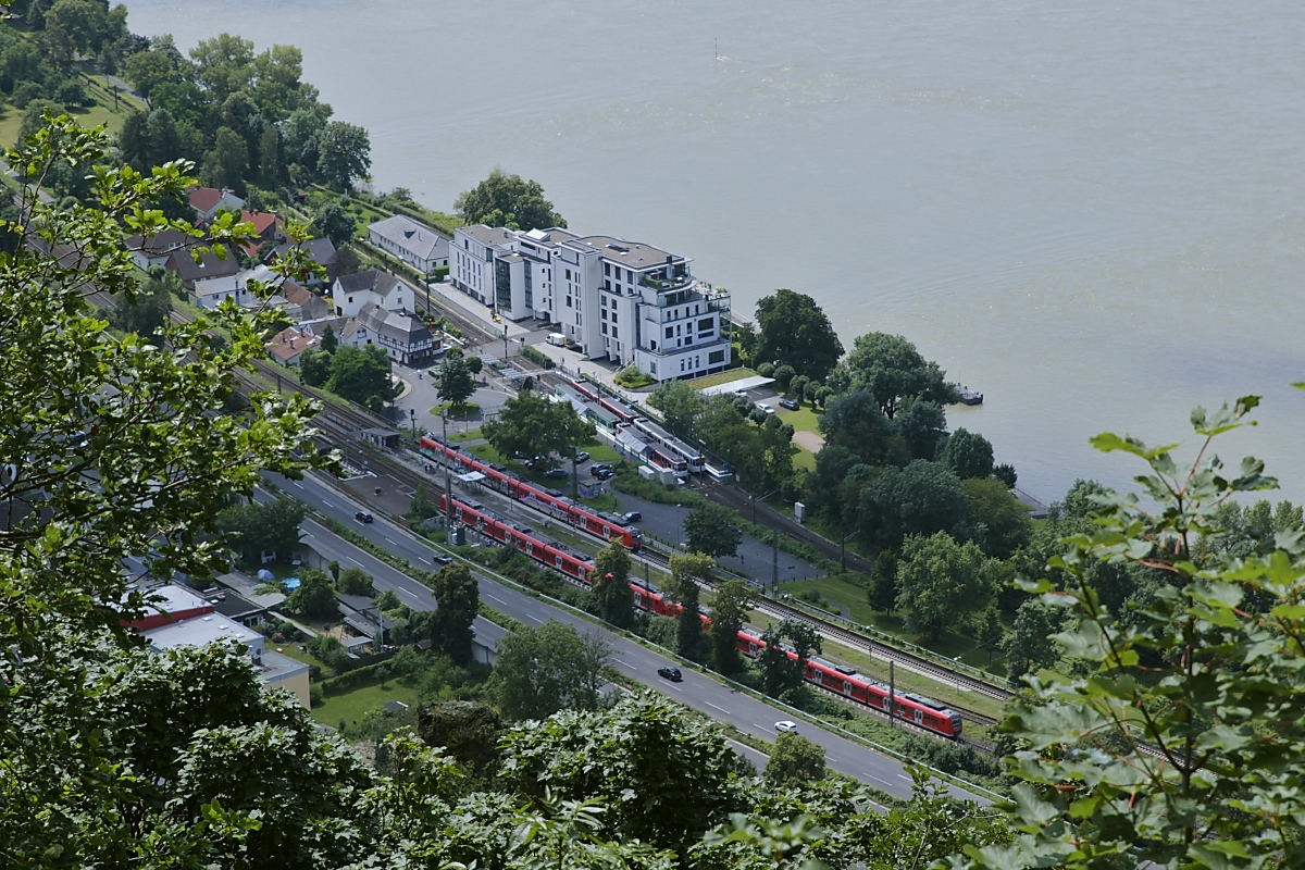 Blick vom Drachenfels hinunter auf den Bahnhof Rhöndorf an der rechtsrheinischen Bahnstrecke am 22.07.2021: Dort treffen sich zwei Züge der RB 27 (Koblenz - Mönchengladbach), bei genauerem Hinsehen entdeckt man auch noch zwei Stadtbahnzüge der Linie 66 (Siegburg - Bad Honnef)
