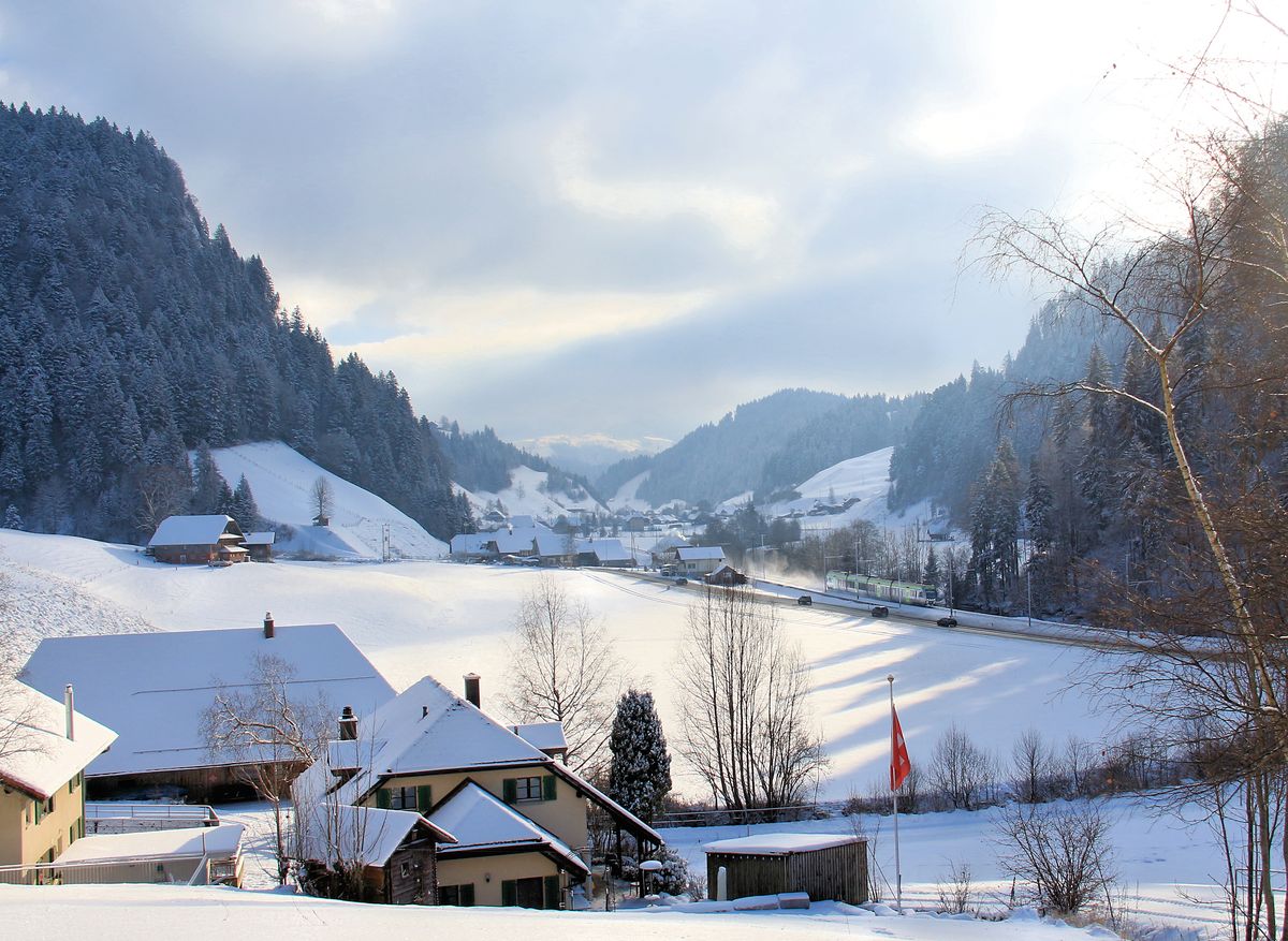 Blick ins  Ausland , von wo sich ein BLS  Lötschberger  aus Luzern nähert. Die Bauernhäuser vorne dürften noch im Kanton Bern sein, die hinten in anderem Stil mit dem kleinen Dach an der vorderen Hauswand im Kanton Luzern. Zwischen Trubschachen (BE) und Wiggen (LU), 6.Januar 2017. 