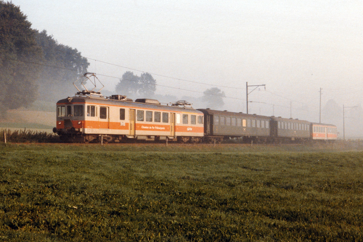 CHEMINS DE FER FRIBOURGEOIS GRUYERE - FRIBOURG - MORAT/GFM.
ERINNERUNGEN AN DIE ORANGE EPOCHE DER STRECKE FRIBOURG - MORAT - ANET.
Am Ende der 70er-Jahre wurde der Anstrich grün/crème  durch orange/silber abgelöst.
Das neue Erscheinungsbild verleihte  dem älteren Rollmaterial ein moderneres Aussehen.
Neblige Stimmung bei Cressier am 5. Oktober 1986. Besondere Beachtung gilt den beiden grünen ex SBB Personenwagen. Sie dienten der GFM als eiserne Reserve sowie als Verstärkungswagen und waren dadurch nur sehr selten im Einsatz zu sehen. 
Foto: Walter Ruetsch  