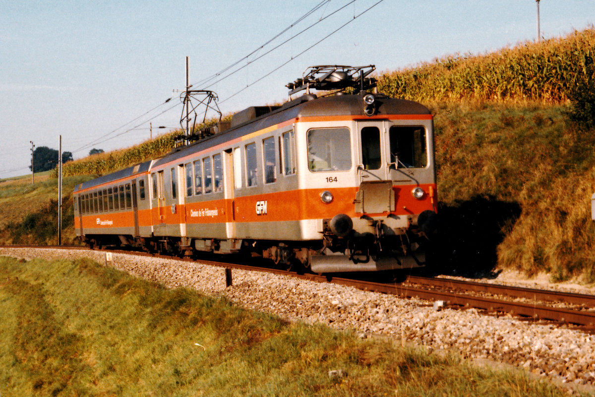 CHEMINS DE FER FRIBOURGEOIS GRUYERE - FRIBOURG - MORAT/GFM.
ERINNERUNGEN AN DIE ORANGE EPOCHE DER STRECKE FRIBOURG - MORAT - ANET.
Am Ende der 70er-Jahre wurde der Anstrich grün/crème  durch orange/silber abgelöst.
Das neue Erscheinungsbild verleihte  dem älteren Rollmaterial ein moderneres Aussehen.
Der ABDe 4/4 164 mit einem Personenwagen der neusten Generation als Regionalzug vor Pensier am 6. September 1986.
Foto: Walter Ruetsch   