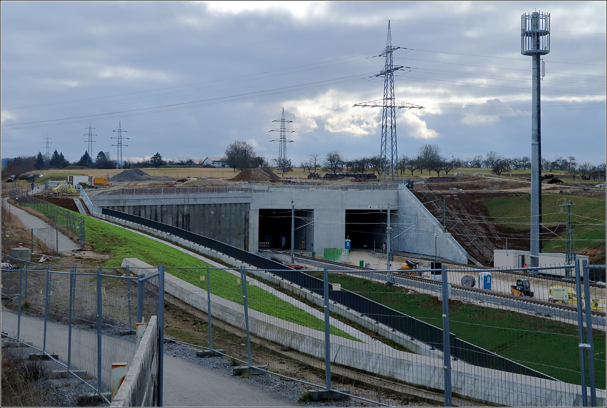 Da wo sich - 

... Stuttgart 21 und die NBS nach Ulm die Hand geben: Wendlingen. 

Ein Blick in die andere Richtung zum Portal des ca. 8,2 km langen Albvorlandtunnels, dessen zwei Röhren mit einer Tunnelbohrmaschine aufgefahren werden. Südöstlich von Kirchheim/Teck befindet sich das andere Tunnelportal.
Im Bild nicht sichtbar das Tunnelportal der Güterzuganbindung. Dieses befindet sich in tiefer Lage im Bereich der Bohrpfahlwand links. Die Güterzuganbindung und die Neubaustrecke nach Ulm befindet sich seit 31.01.2022 und Testfahrten können durchgeführt werden.

Letzten Sommer war ich schon mal hier. Da war der Rad- und Fußweg als Durchgangsweg offiziell gesperrt, aber von den Feldwegen von hinten her dennoch offen zugänglich. Die damals gemachten Fotos habe ich vorsichtshalber damals nicht veröffentlicht.

04.02.2022 (M)