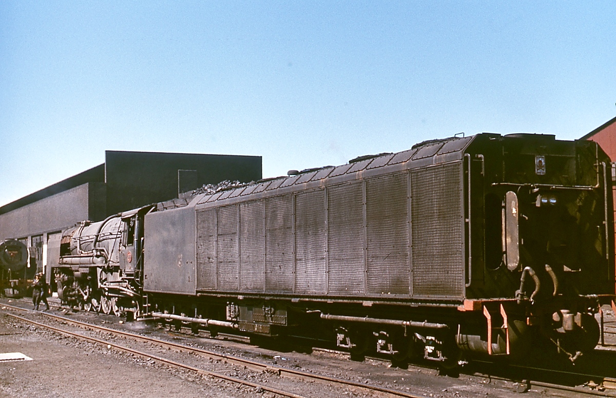 Dampf und Diesel in der Karoo: Blick auf den Tender einer Kondenslokomotive der Class 25 im November 1976 im Depot De Aar. Die Karoo, eine Halbwüste, nimmt etwa 1/3 der Fläche Südafrikas ein. Die Wasserversorgung der Dampflokomotiven bereitete hier stets Probleme. Um Abhilfe zu schaffen, bestellte die SAR bei Henschel und North British 90 Kondenslokomotiven, bei denen der Abdampf dem Tender wieder zugeführt wird. Im Vergleich mit konventionellen Dampflokomotiven konnte der Wasserverbrauch um etwa 85% gesenkt werden. Bis auf 3 wurden die 1954/54 in Dienst gestellten Kondensmaschinen ab 1974 wegen der fortschreitenden Elektrifizierung und Verdieselung zu 25 NC (=  non condens ) umgebaut.