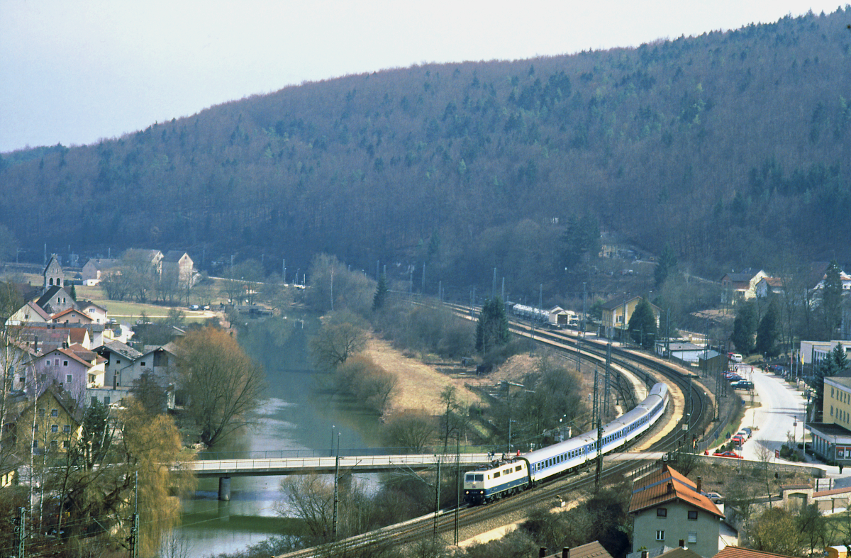 Das Foto zeigt den Bahnhof Solnhofen am 7. April 1996. Der Fluß ist die Altmühl. Eine Lok 111 durchfährt den Bahnhof mit einem Interregio München - Würzburg. Gerade hat sie den Bahnübergang bei Kilometer 125,5 passiert. Im Hintergrund sind Zementwaggons auf den Gütergleisen des Bahnhofs zu erkennen. Interregio, Gütergleise und Zementwaggons sind längst vergangen, aber der Zugverkehr ist heute dichter denn je, es dominieren Güterzüge.