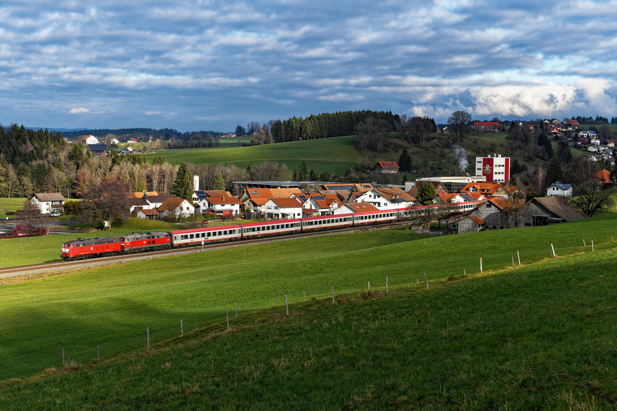 Das Fotografenglück für dieses Jahr habe ich bei diesem Bild vermutlich schon komplett ausgeschöpft. Am 11. Januar 2020 gab sich eine weitere Leihlok vor dem für eine kurze Periode wieder zwischen Lindau und Stuttgart in Verkehr gesetzten IC  Bodensee  die Ehre. Die orientrote 218 402 von Railsystems brachte den IC 119 gemeinsam mit der Ulmer 218 491 umleitungsbedingt über die Allgäubahn zu seinem Zwischenziel. Leider zeigte sich der Tag bewölkt und regnerisch. Trotzdem war ich im Westallgäu unterwegs, die freie Zeit sollte zumindest zur Erkundung von Fotostellen verendet werden. Das Motiv mit dem Blick auf die Ortschaft Meckatz samt Brauerei hatte ich bisher nicht umgesetzt und stand noch auf meiner to-do Liste. Bei Durchfahrt des Zuges riss die Bewölkung jedoch plötzlich auf und ermöglichte diese  Theaterbeleuchtung  auf diese aufgrund Bespannung und Umleitung einmalige Leistung auf der Kursbuchstrecke 970. 