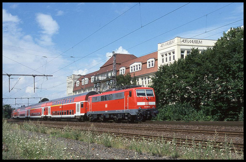 DB 111089 fährt hier am 4.8.2002 um 12.28 Uhr mit ihrem Dosto aus Richtung Osnabrück kommend in den Bahnhof Melle ein. Dabei passiert sie das Meller Automuseum, welches früher eine alte Möbelfabrik war.