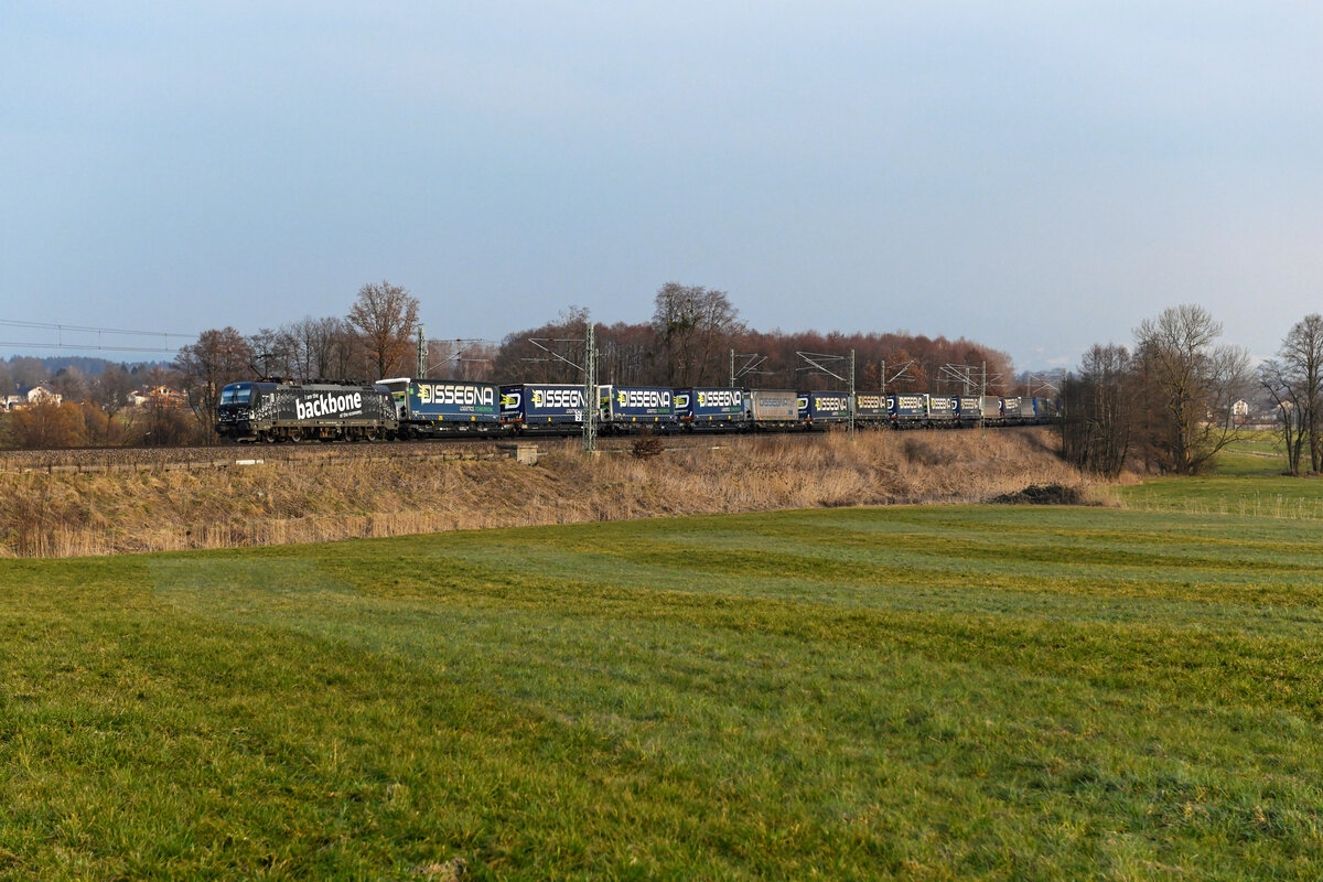 Der 04. März 2023 zeigte sich am späteren Nachmittag im bayerischen Voralpenland sehr dunstig. Normalerweise hätte ich unter solchen Bedingungen längst die Kamera weggepackt und mich auf den Heimweg begeben. Allerdings sollte von Kufstein her die 193 365 mit einem KLV-Zug westwärts fahren. Das auffällige Design der Lok mit dem Rückgrat-Motiv soll die Bedeutung der Bahn für die Wirtschaft und den Warenverkehr hervorheben. Normalerweise verkehren die Vectrons von DB Cargo mit Nummern ab 360 aufwärts zwischen Polen und den Benelux-Ländern. Einsätze in Bayern sind extrem rar. Daher vertrieben sich ein Hobbykollege und ich die Zeit mit Gesprächen, bis dann tatsächlich die  Backbone  mit einem gut beladenen KLV-Zug bei Rann durchfuhr. 