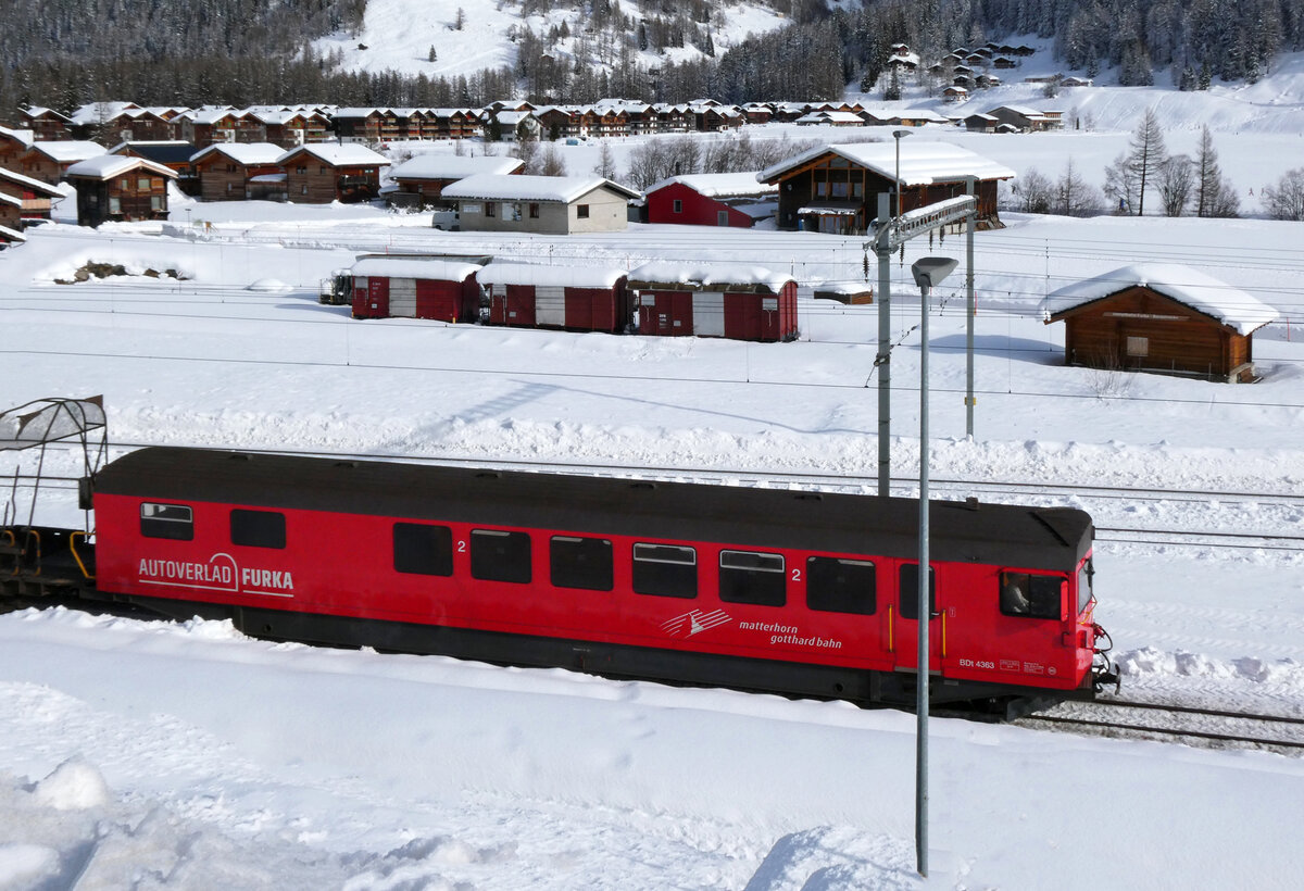 Der Autozug von Realp durch den Furkabasistunnel fährt mit dem Steuerwagen voraus in Oberwald ein. Im Hintergrund sind drei eingeschneite Güterwagen sowie das Stationsgebäude der DFB (Dampfbahn Furkabergstrecke) zu sehen. Oberwald, 13.02.2026