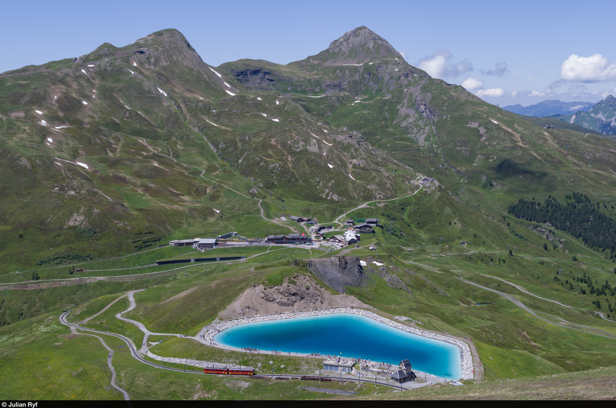 Der BDhe 2/4 210 der Jungfraubahn fährt am 24. Juni 2015 am Speichersee Fallboden vorbei talwärts in Richtung Kleine Scheidegg, deren Bahnhof weiter unten zu erkennen ist. Von dort geht es mit der Wengernalpbahn weiter: rechts nach Grindelwald, links nach Wengen-Lauterbrunnen. An beiden Orten besteht Anschluss an die Berner Oberland Bahn nach Interlaken Ost - welchen Weg man nimmt spielt auch zeitlich keine Rolle. 
Über der Kleinen Scheidegg sind links das Lauberhorn (bekannt durch die Ski-Weltcup-Abfahrt) und rechts der Tschuggen zu sehen.