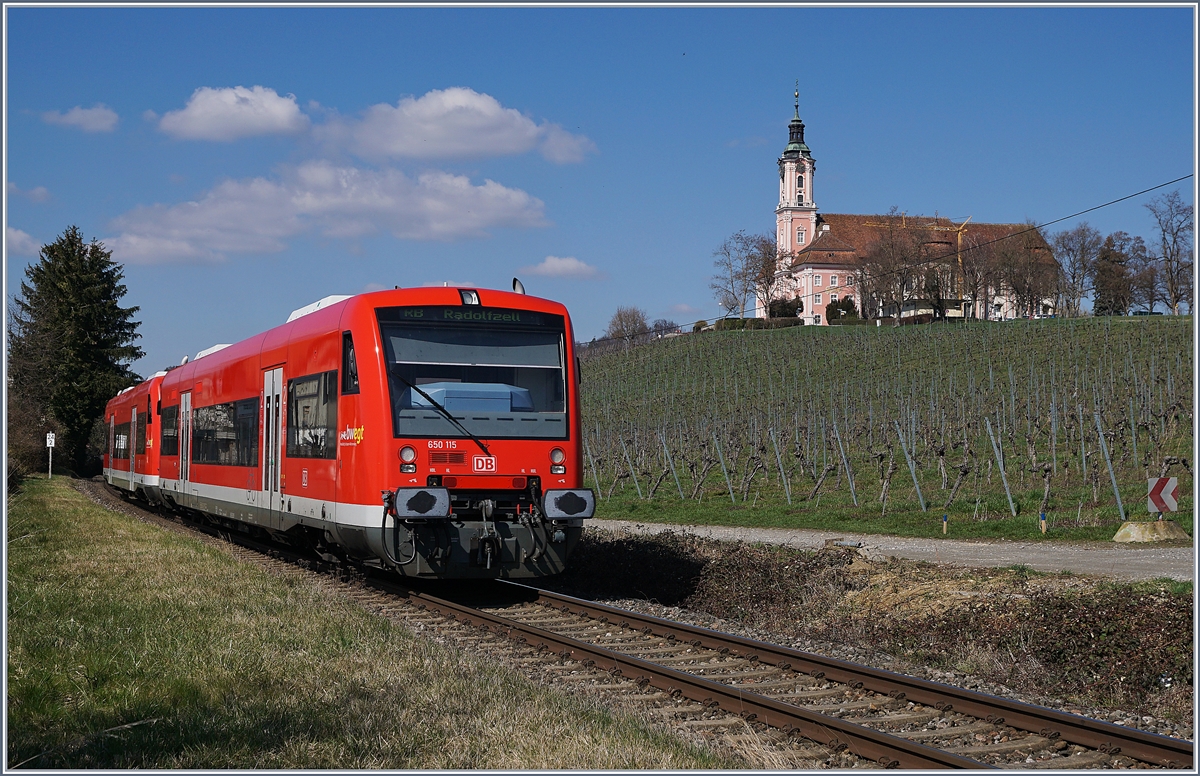 Der DB 650 115 und ein weiterer sind als RB 22766 von Friedrichhafen nach Radolfzell kurz vor der ehemaligne Station Birnau-Maurach unterwegs; rechts im Bild die Barockkirche Birnau. 


20. März 2019
