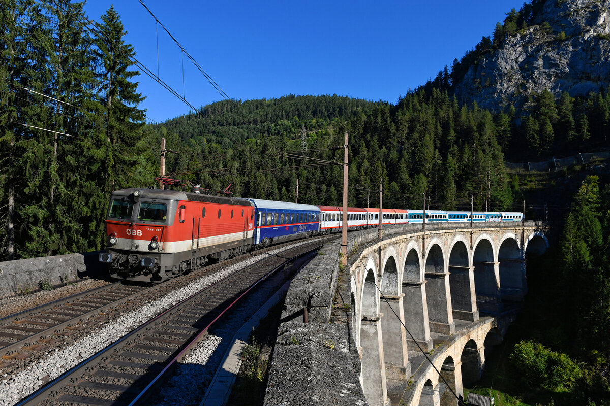 Der EC 151  Emona  von Wien HBF nach Ljubljana ist noch ein klassischer internationaler Fernzug. Bis Ende 2021 war dieser EuroCity eine der wenigen verbliebenen hochwertigen Reisezugleistungen für die Baureihe 1144. Am 25. September 2021 konnte ich mit der 1144.027 auch eine besondere Vertreterin dieser Baureihe vor dem besagten EC 151 auf dem Kalte Rinne Viadukt am Semmering fotografieren. Dieses Bauwerk der ältesten Gebirgsbahn Europas wurde bereits im Jahre 1853 fertiggestellt und zählt zum Weltkulturerbe. Was ist nun so besonders an der 1144.027? Auf dieser Aufnahme erkennt man es gut: Auf einer Seite des Lokkastens weisen die Bullaugen eine unterschiedliche Höhe auf. Vermutlich ein Fertigungsfehler beim Ausstanzen des Blechs. Es wird sich aber vermutlich nicht mehr klären, was 1978 in den Werkshallen von SGP damit bezweckt wurde, oder ob das eine  bsoffene Gschicht  war....